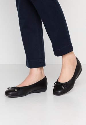 Persoon die zwarte leren ballerina's met kleine strikjes draagt, gecombineerd met donkerblauwe cropped broeken, staand op een lichte ondergrond.