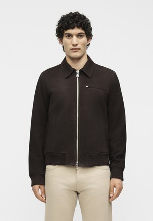 Jeune homme aux cheveux bouclés portant une veste marron foncé zippée et un pantalon beige, debout devant un fond blanc uni.