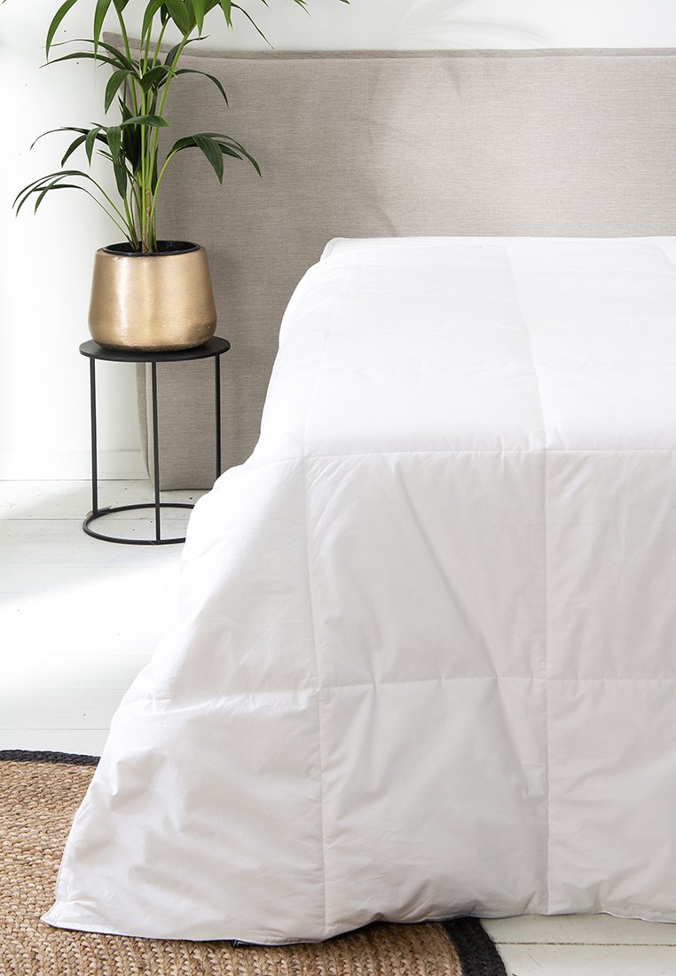 Piumone bianco trapuntato parzialmente coprente un letto, accompagnato da una pianta in vaso dorato su un tavolino laterale in metallo nero, su uno sfondo neutro.