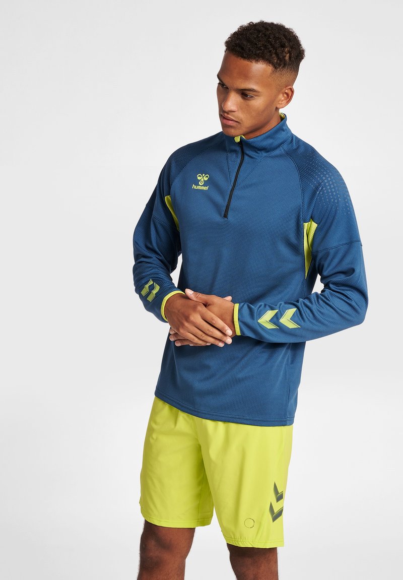 Pullover sportivo blu con accenti gialli, mezzo zip, tessuto testurizzato e logo. Abbinato a pantaloni corti gialli brillanti con motivi.