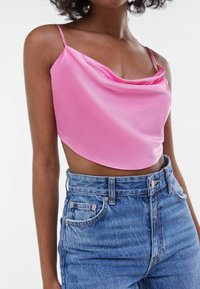 Růžový saténový crop top s drapovaným výstřihem a tenkými ramínky, spárovaný s modrými džínami vysokého pasu, které mají přední kapsy a zapínání na knoflík.
