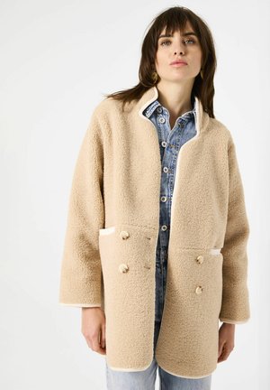 Abrigo de sherpa beige con un acabado texturizado, cierre de doble botonadura y ribete blanco en contraste. Presenta dos bolsillos delanteros.