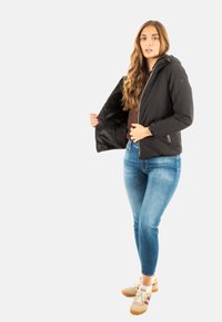 Giacca nera con zip e cappuccio, con una morbida fodera interna e polsini elasticizzati, abbinata a jeans skinny blu e sneakers beige.