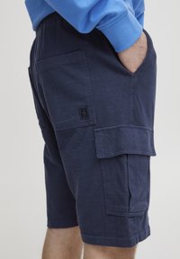 Tamsiai mėlyni medvilniniai krovininiai šortai su šoninėmis kišenėmis ir maža juoda prekės ženklo etikete. Audinys minkštas, lygios tekstūros ir pritaikyto silueto.