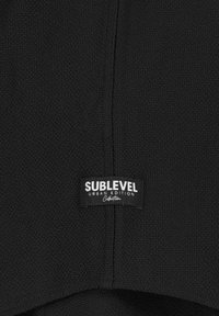 Schwarzes gewebtes Stoffetikett mit dem Text "SUBLEVEL URBAN EDITION" in Weiß, mit strukturiertem Finish und Nähdetails.