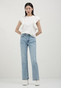 T-shirt bianca in cotone con maniche a volant, abbinata a jeans a gamba dritta blu chiaro e sneakers bianche; design minimalista e casual.