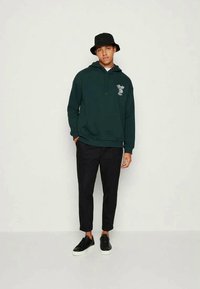 Πράσινη hoodie με γραφικό σχέδιο στο στήθος, συνδυασμένη με μαύρο tapered παντελόνα και μαύρα αθλητικά παπούτσια. Το μοντέλο φοράει μαύρο καπέλο bucket.