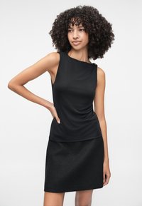 Μαύρη αμάνικη μπλούζα με σχέδιο που πέφτει και διακριτική λεπτομέρεια στον ώμο, συνδυασμένη με μια υφασμάτινη μαύρη φούστα. Λείο φινίρισμα υφάσματος.