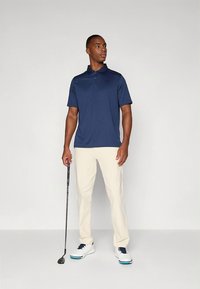 Mörkblå pikétröja med korta ärmar, ljusbeiga byxor och vita sportskor. Personen håller i en golfklubba.