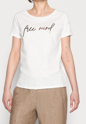 Person trägt ein weißes T-Shirt mit dem Schriftzug "free mind" und hellbraune Hose, steht vor einem einfarbigen Hintergrund.