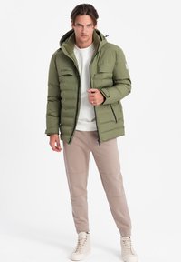 Olivově zelená péřová bunda s kapucí, zipovým zapínáním a několika kapsami. Nositá přes krémovou mikinu a béžové zúžené kalhoty.