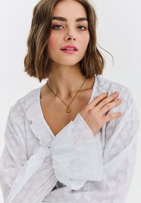 Młoda kobieta o brązowych włosach do ramion, ubrana w białą teksturowaną bluzkę i złoty naszyjnik z wisiorkiem w kształcie kompasu, z ręką spoczywającą na klatce piersiowej.
