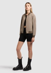 Femme debout portant une veste bomber taupe, un crop top noir, des shorts de cycliste noirs et des bottes noires épaisses mi-mollet sur un fond gris.