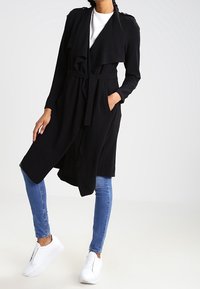 Manteau portefeuille noir en tissu doux, avec un devant drapé, des manches longues et une ceinture à nouer, porté avec un jean bleu et des baskets blanches.