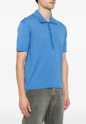 Man draagt een blauw polohemd met korte mouwen en zwarte knopen en vervaagde grijze spijkerbroek, staand tegen een witte achtergrond.