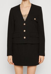 Černý blazer s výstřihem do V a malou přední kapsou, se zlatými knoflíky a texturovanou tkaninou, kombinovaný s topem s leopardím vzorem.