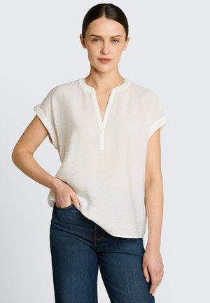 Femme aux cheveux foncés tirés en arrière, portant un chemisier blanc à manches courtes et un jean bleu foncé, debout avec une main dans la poche.
