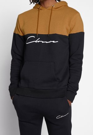 Mann trägt einen schwarz-braunen Hoodie mit weißem Schriftzug „Closure“ und passende schwarze Hose mit Schriftzug „Closure“ am Oberschenkel.