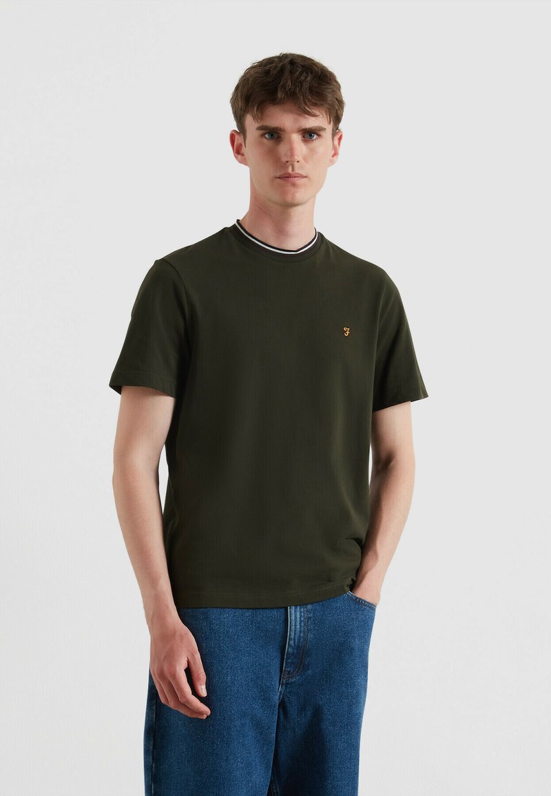 T-shirt a maniche corte di color verde oliva con colletto rotondo a righe e un piccolo logo sul petto. Indossato con jeans in denim blu.
