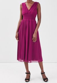 Robe fuchsia plissée avec un col en V et un corsage ajusté, s'étendant en dessous du genou. Associée à des sandales à talons noires à lanières.