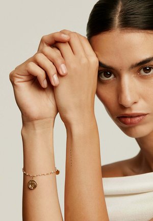 Pulsera de oro con un charm redondo, pequeñas piedras y delicada cadena. El modelo tiene la piel clara, con las manos entrelazadas, mostrando un suave esmalte de uñas.