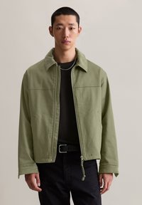 Grüne, Reißverschlussjacke mit kragenlosem Ausschnitt, aus weichem Stoff gefertigt, mit einer vorderen Naht und elastischem Zug am Saum. Über einem schwarzen Shirt getragen.