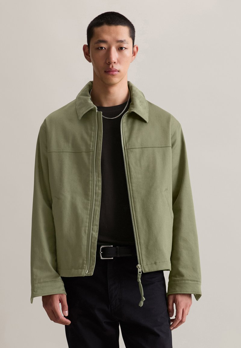 Grüne, Reißverschlussjacke mit kragenlosem Ausschnitt, aus weichem Stoff gefertigt, mit einer vorderen Naht und elastischem Zug am Saum. Über einem schwarzen Shirt getragen.