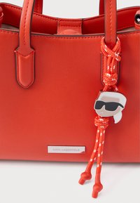Punainen nahkainen käsilaukku, jossa on kaksinkertaiset kahvat, teksturoitu pinta ja irrotettava koriste, jossa valkohiuksinen hahmo aurinkolaseissa, kiinnitetty oranssilla narulla.