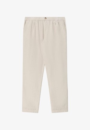 Mann mit dunklem Haar, der ein senfgelbes T-Shirt mit hochgekrempelten Ärmeln, hellbeige Hose und braune Slipper trägt, steht mit Händen in den Taschen.