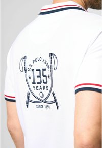 Hvid poloskjorte med broderet design, der viser "U.S. Polo Assn. 135 År" og krydsede maletter. Kantet med røde og marineblå accenter.