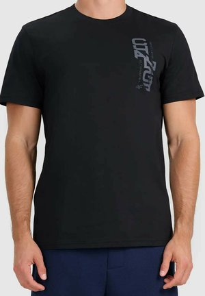 Mężczyzna w gładkiej czarnej koszulce z krótkim rękawem z ciemnym pionowym napisem na górnej lewej stronie klatki piersiowej, w połączeniu z ciemnoniebieskimi spodniami.