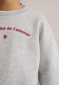 Sivkasta srajca s strukturovanim ovratnikom in bardo rdečim besedilom "Amour de l'amour" z majhnim srcem spodaj. Mehka, teksturirana tkanina.
