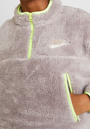Zbliżenie osoby noszącej jasnoszary, miękki pulower Nike z neonowożółtym zamkiem błyskawicznym i ręką w kieszeni na zamek.