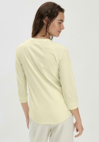 Hellgelbes Shirt mit drei Viertel-Ärmeln, rundem Halsausschnitt, weichem Stoff und abgerundetem Saum, kombiniert mit hellen Hosen.