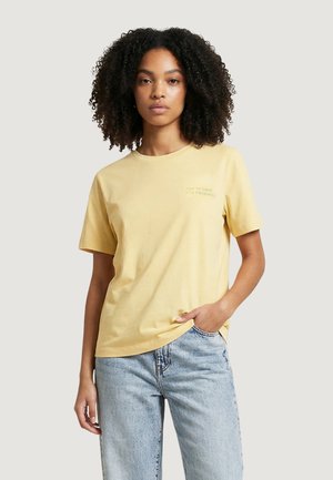 Giovane donna con capelli ricci che indossa una maglietta a maniche corte giallo pallido e jeans blu chiaro, in piedi con una mano in tasca.