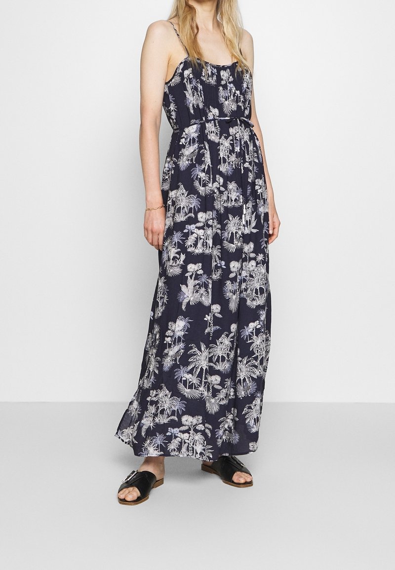 Femme portant une robe maxi bleu marine à imprimé floral blanc avec des bretelles fines, associée à des sandales noires ouvertes à l'avant.