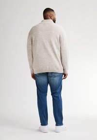 Beige pletený svetr s vysokým límcem a žebrovanými detaily na ramenních švech, spárovaný s modrými džíny a bílými teniskami. Jednoduchý, strukturovaný design.