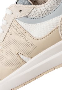 Sneaker beige con tomaia in pelle liscia e mesh, con dettagli perforati e una suola in gomma bianca. Dettagli azzurri leggeri sulla linguetta.