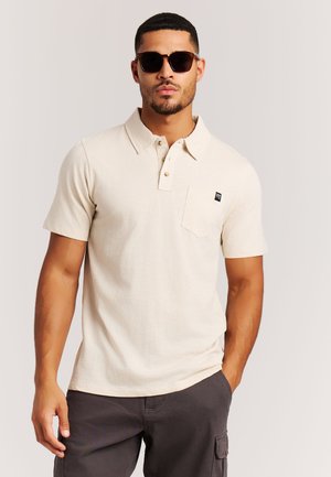 Man draagt een beige polo met korte mouwen en borstzak, en donkere zonnebril, met één hand in de zak van donkere cargobroek.