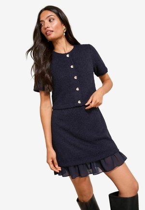 Marineblaues Kleid mit kurzen Ärmeln, strukturiertem Stoff, einer Reihe goldfarbener Knöpfe, geschichtetem transparentem Rüschensaum, kombiniert mit kniehohen schwarzen Stiefeln.