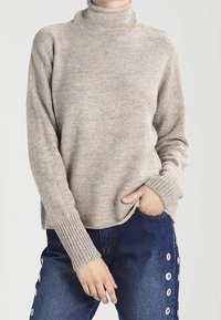 Person, der einen beigen Rollkragenpullover mit langen Rippbündchen und blaue Jeans mit weißen Ösen-Details an den Seiten trägt.