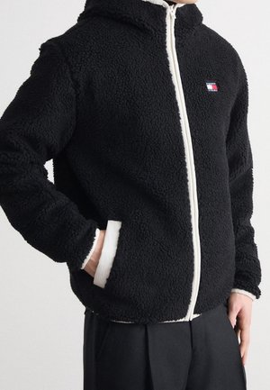 Personne portant une veste polaire noire zippée avec des bordures blanches et des poches, avec un petit logo Tommy Jeans sur la poitrine.