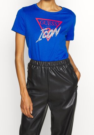 Osoba nosząca niebieski t-shirt Guess USA z napisem „ICON” z cyrkonii, połączony z czarnymi, wysokim stanem, sztucznymi skórzanymi spodniami z kieszeniami.