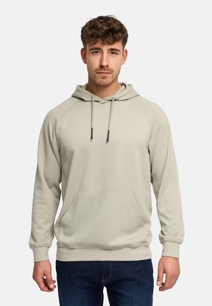 Junger Mann mit lockigem Haar, der einen hellbeigen Hoodie mit Fronttasche und dunkelblaue Jeans trägt, steht vor einem einfarbigen Hintergrund.