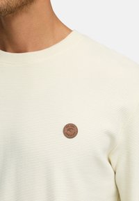 Helle, gerippte Sweatshirt mit rundem Ausschnitt und einem braunen, runden Lederpatch mit erhabenem Text auf der linken Brust.