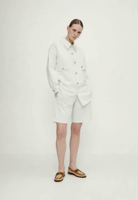 Balta marškinėliai su mygtukais ir dviem priekinėmis kišenėmis, derinami su iki kelių ilgio šortais. Nešiojami su rudais slip-on batais, prieš paprastą foną.