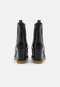 Bottines en cuir noir avec un talon compensé haut, texture lisse, côtés élastiques et semelle beige contrastante. Une languette à l'arrière.