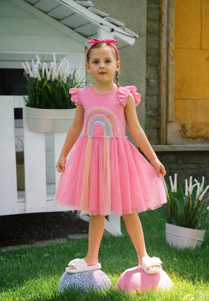 Junges Mädchen in pinkfarbenem Regenbogenkleid und Sandalen steht draußen auf bunten kuppelförmigen Trittsteinen im Gras.