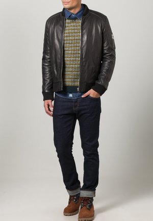 Hombre con chaqueta de cuero negra, suéter de patrón, camisa de mezclilla azul, jeans oscuros y botas marrones, de pie con una mano en el bolsillo.