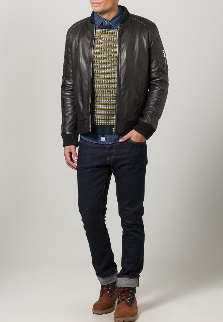 Man in een zwart leren jack, met een patroontrui, een blauw denim shirt, donkere jeans en een paar bruine laarzen, staat met één hand in zijn zak.